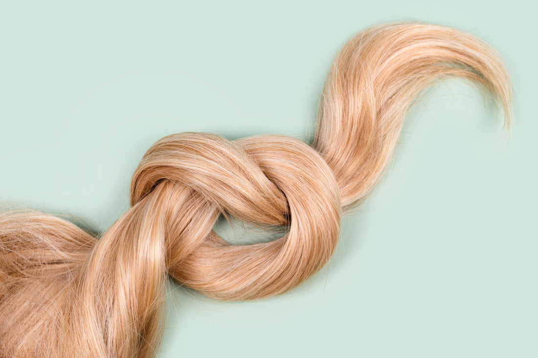 A very long lock of shiny and lustrous blonde hair tied into a knot. It rests on a pale green background.