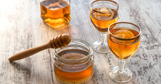 A honey dipper rests against the side of a jar of honey next to two glasses filled with an amber-colored drink.