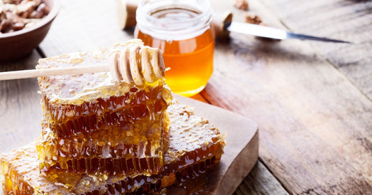 Stacked honeycomb sits on a piece of wood. A honey dipper lies on the honeycomb with a jar of honey behind it.
