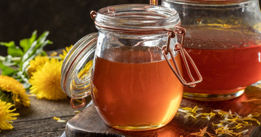 Two glass jars full of honey sitting on a wooden board that rests on a table next to yellow dandelions and scattered petals.