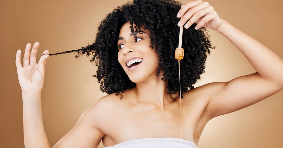 A woman holds a strand of her curly hair out to the side while holding a wooden honey dipper with honey on it.