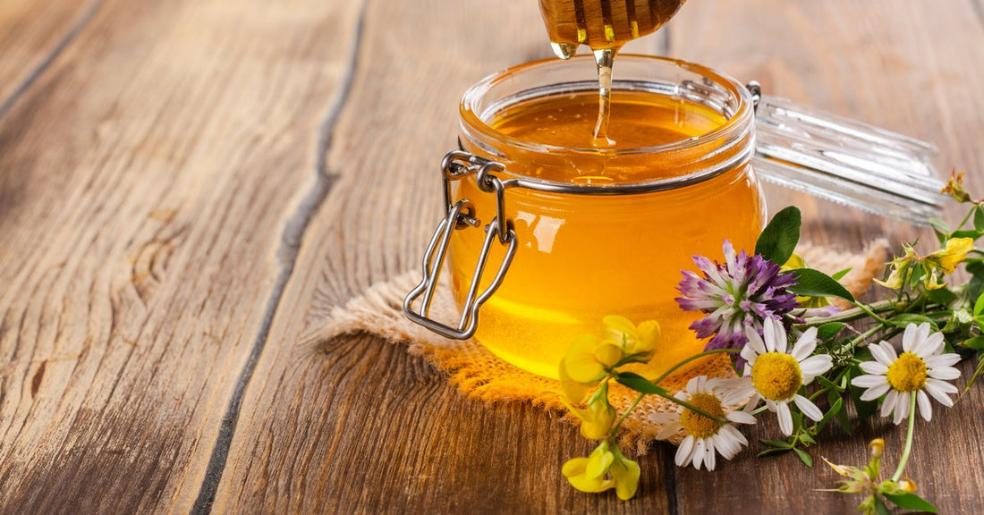 A wooden honey dipper drips into a glass container full of honey. A variety of flowers sit on the table with the honey.