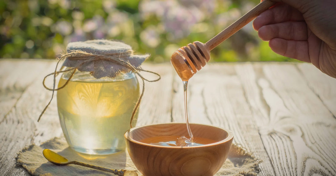 Honey drips into a wooden bowl from a honey dipper held in the air. It sits next to a jar of honey on a table outside.