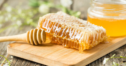  A honeycomb and a jar of honey sit on a wooden cutting board next to a honey dipper. Small white flowers are scattered about.