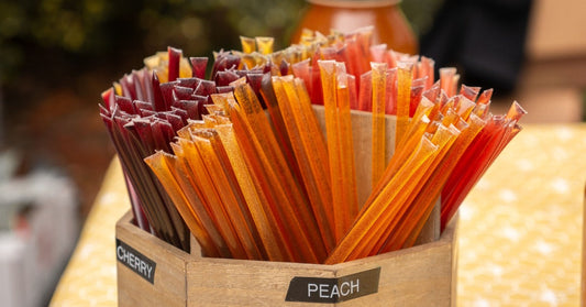 A wooden box filled with different colored honey sticks. The box has labels that say "CHERRY" and "PEACH."