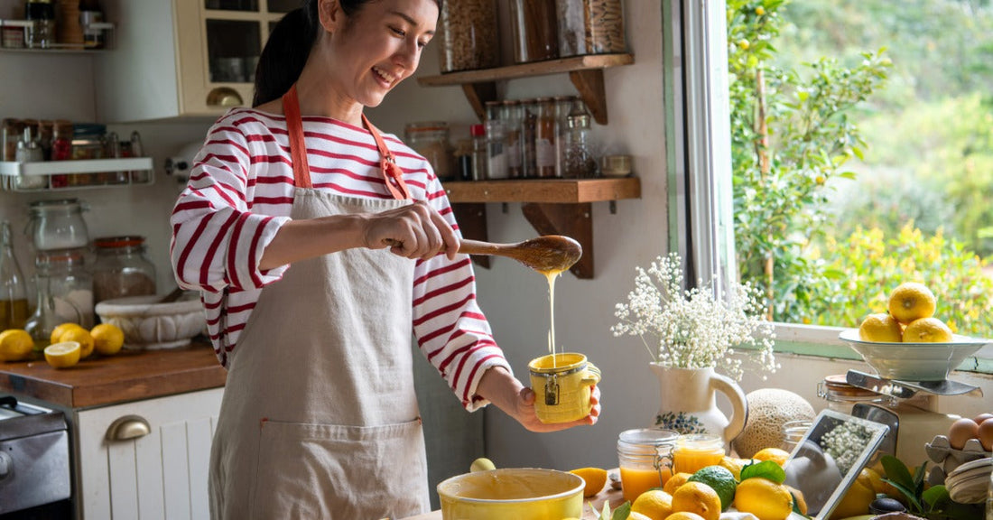  Person in a striped shirt and apron pours homemade lemon curd into a glass jar in a bright kitchen filled with lemons.