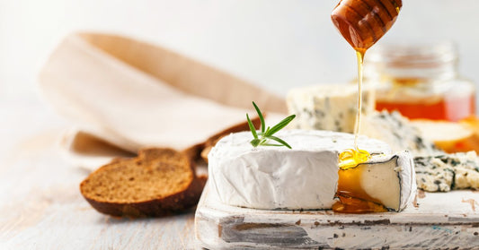Golden honey drips from a wooden dipper onto a wheel of soft white cheese served with bread and blue cheese.
