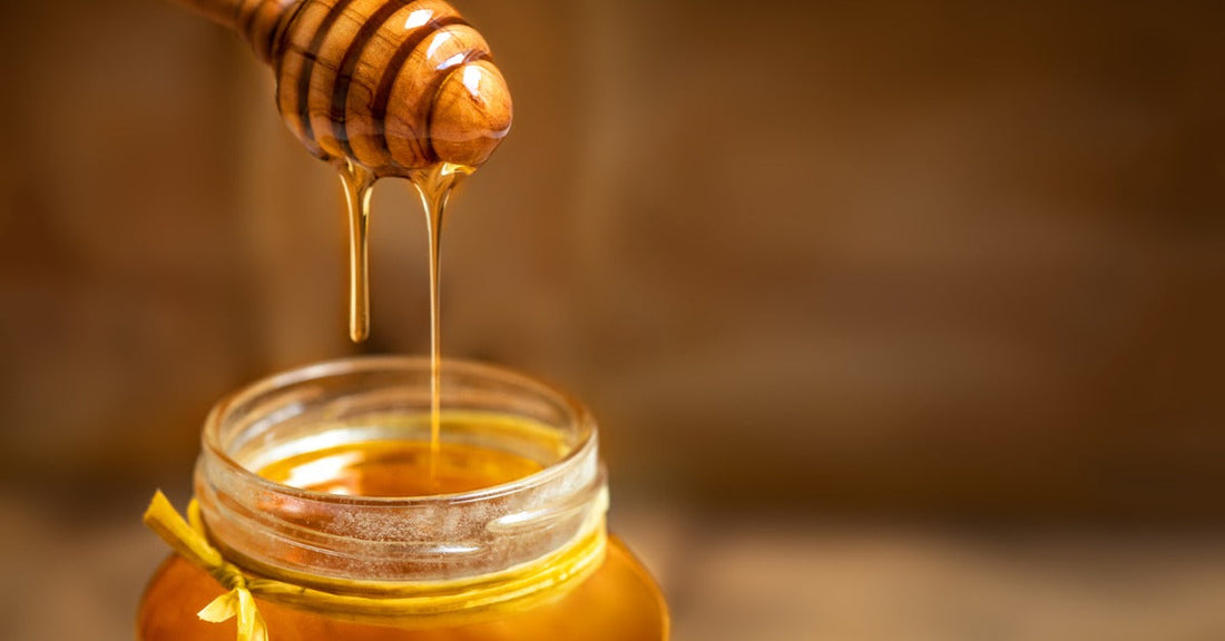 Honey drips from a wooden dipper into a glass jar, with warm golden light and a blurred brown background.