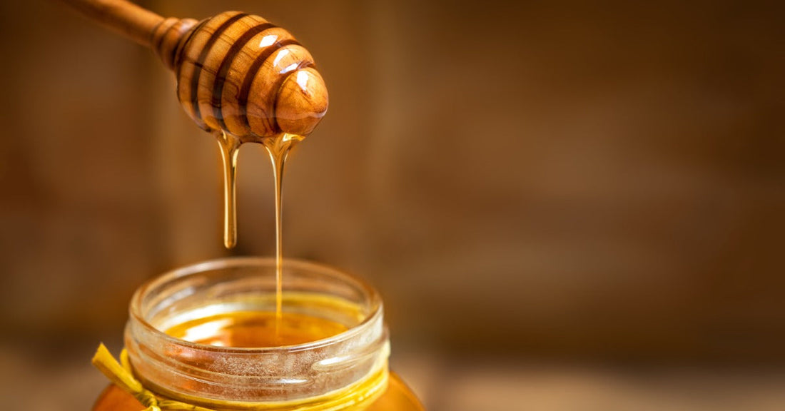 A honey dipper coated in golden honey hangs above a glass jar as thick streams fall into the opening.
