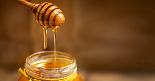 A honey dipper coated in golden honey hangs above a glass jar as thick streams fall into the opening.