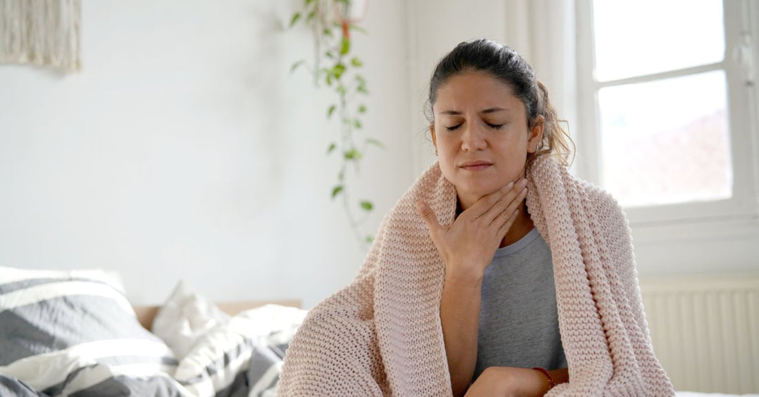 A woman wrapped in a pink blanket sits up in bed. She is grimacing with her eyes closed and touching her throat.