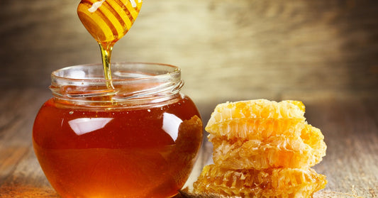 A honey dipper drips with honey over a glass jar of raw honey. To the right is a stack of three pieces of honeycomb.