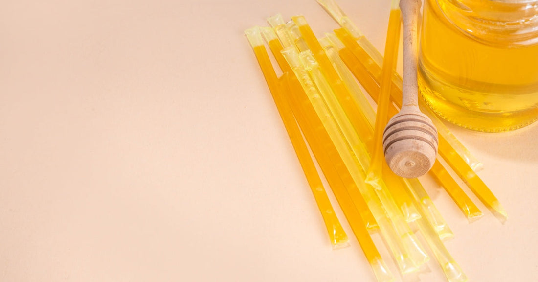 A honey dipper lies on top of a pile of honey sticks next to a jar full of golden honey set against a pink background.