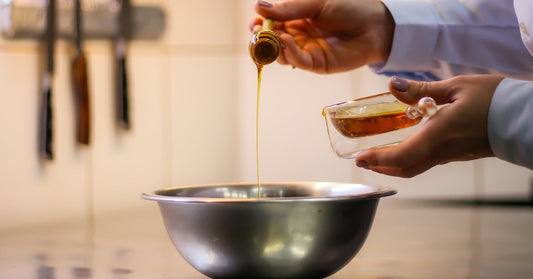 A person wearing a chef's jacket holds a honey dipper over a stainless-steel bowl. They hold a glass jar in their other hand.