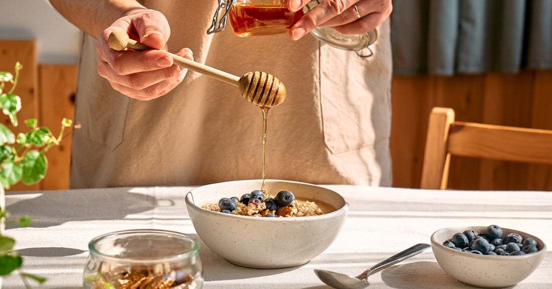 A person wearing an apron uses a honey dipper to drizzle honey over a bowl of oatmeal with blueberries.