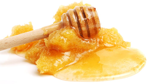 A wooden honey wand lies on a pile of crystallized honey against a white background. Liquid honey seeps from the mound.