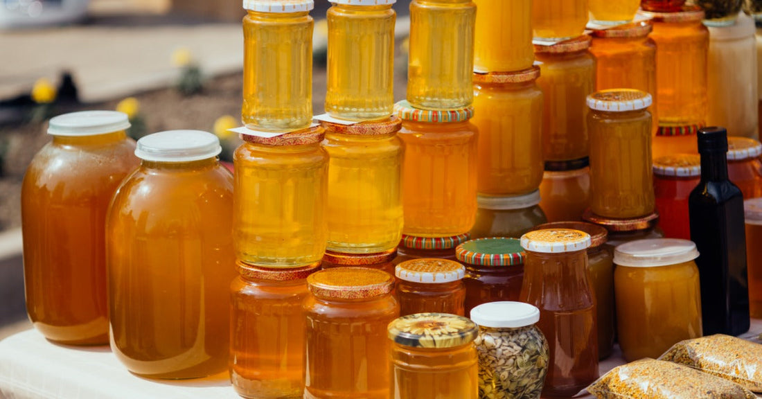 Rows of glass jars filled with golden honey sit on a table outdoors, showing a variety of sizes, shades, and textures.