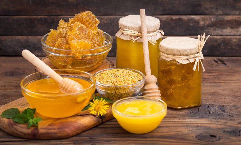 Two jars and four bowls full of bee-related products, such as honey, pollen, and jelly, sit on a wooden table.