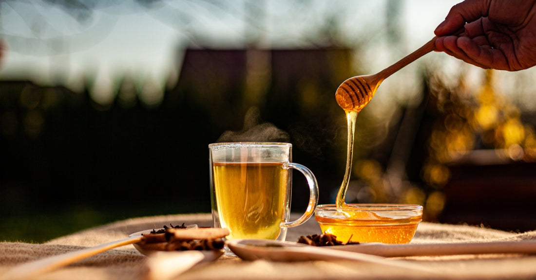 A clear mug filled with tea stands beside a small shallow bowl filled with honey that a honey dripper is dripping honey into.