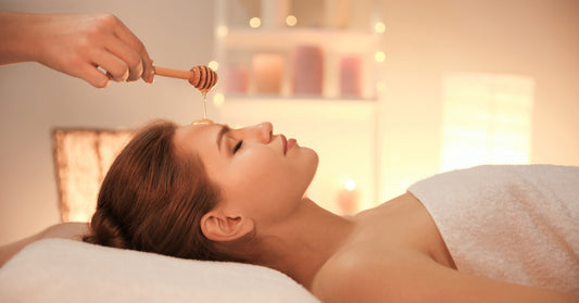 A woman in a towel has her eyes closed and is laying down on a massage table as the masseuse applies a honey mask.