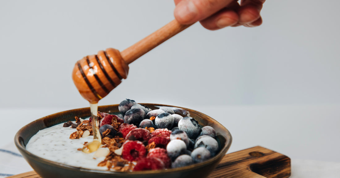 A woman's hand holding a honey dripper, drizzling honey across a bowl full of fruit, grains, and yogurt.