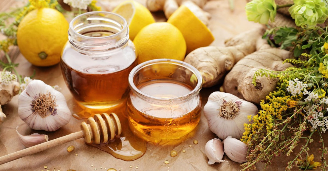 Two jars of honey are surrounded by garlic, lemon, herbs, ginger, and other elements for infusion.