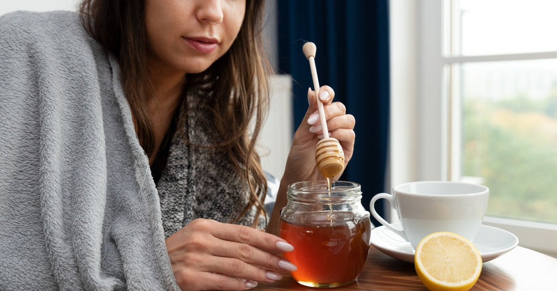 A woman sitting by a window with a blanket wrapped around her shoulders pulls a honey dipper up from a jar of honey.